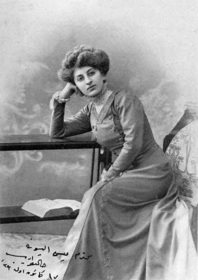 This is a formally posed almost
full-body seated photo of the writer. She is leaning her left elbow
on a two-tiered table with an open book on the lower shelf, with her closed hand
against her cheek. She is wearing a long sleeved dress, and her hair is up but not
covered. There is a hand-written inscription in Arabic script in the
lower left of the photo.